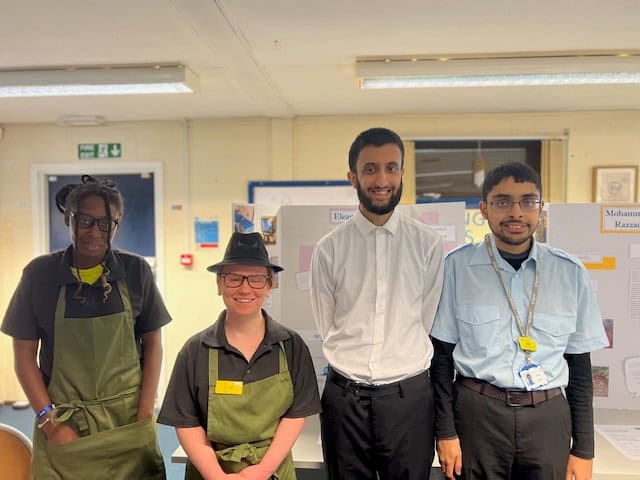 Four people lined up, two females and two males. The females are wearing hats and aprons whilst the males are wearing shirts and trousers.